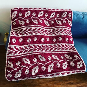 May include: A red and white knitted throw blanket with a geometric pattern. The blanket is folded and placed on a blue couch.