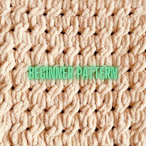 May include: A close-up of a beige knitted fabric with a woven pattern. The fabric has a soft, fluffy texture and is made with a chunky yarn. The text "BEGINNER PATTERN" is printed on the fabric.