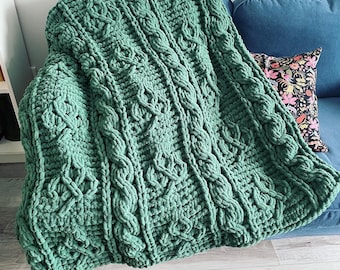 May include: A large, hand-knitted blanket in a soft, muted green color. The blanket features a cable-knit design with vertical and horizontal patterns. It is draped over a blue sofa, with a decorative pillow visible.