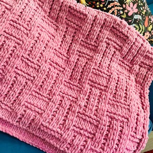 Peut inclure: Une couverture rose tricotée à la main avec un motif en forme de panier tressé. La couverture est faite de fil épais et moelleux, créant un aspect douillet et texturé. La couverture est pliée sur une surface bleue, avec un coussin à motifs floraux en arrière-plan.