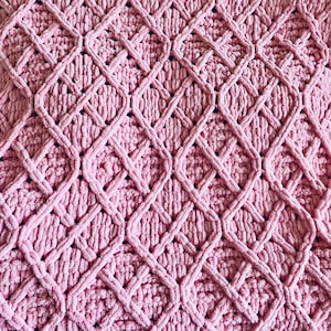 Pode incluir: Um tecido de crochê rosa claro com um padrão de losangos em relevo. A superfície é composta por laçadas grossas de fio felpudo que criam um efeito de grade texturizado e tridimensional em todo o tecido.