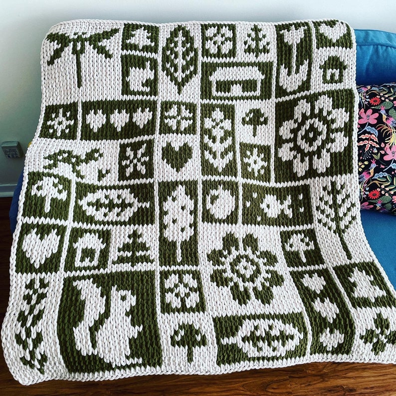 May include: A knitted blanket with a repeating pattern of green and white squares. Each square features a different nature-themed design, including trees, hearts, and flowers. The blanket is draped over a blue couch.