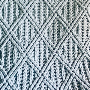 May include: A close-up of a pale blue knitted blanket. The blanket has a textured pattern of interwoven, thick yarn, creating a diamond design. The soft, plush material is visible, suggesting a cosy and comfortable texture.