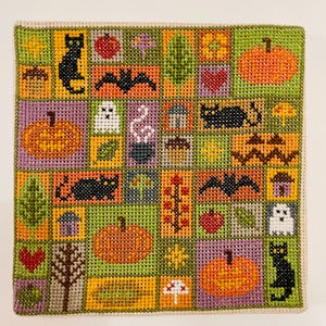 May include: A colorful, square needlepoint featuring Halloween-themed motifs. The design includes pumpkins, bats, black cats, ghosts, and other fall elements. The background is a patchwork of green, orange, and purple squares, creating a festive, handcrafted look.