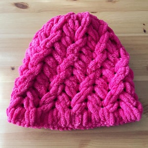 Puede incluir: Un gorro tejido a mano de color rosa brillante. El gorro presenta un diseño de trenza gruesa, creando una apariencia texturizada. El gorro está hecho de un material suave y afelpado, ideal para el invierno.
