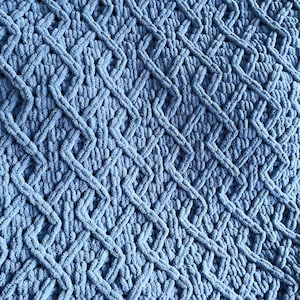 Op de afbeelding: Een blauwe gebreide deken met een ruitpatroon. De deken is gemaakt van dik garen en heeft een zachte, pluche textuur.