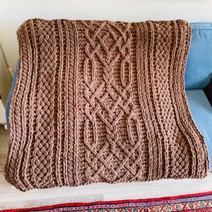 May include: A brown crocheted throw blanket with a cable knit pattern. The blanket is made with thick yarn and has a soft, cozy texture.