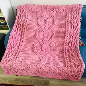 May include: A pink knitted blanket with a cable-knit heart design in the center. The blanket has a textured border and is draped over a blue sofa. The blanket is approximately 60 inches by 50 inches.