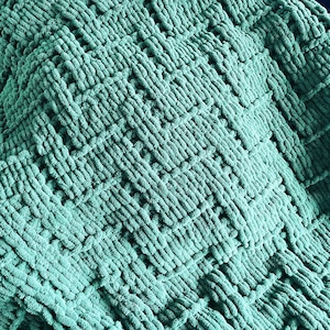 May include: Close-up of a teal-colored, chunky knit blanket. The blanket features a woven pattern with a textured, plush appearance. The weave creates a grid-like design, adding visual interest to the soft, cozy textile. The blanket is a solid color.