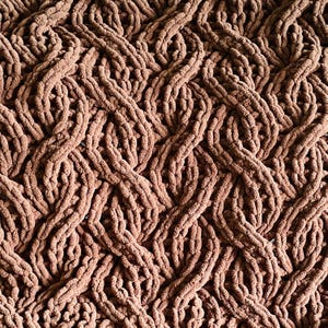 May include: Close-up of a brown, chunky knit blanket with a cable-knit design. The blanket's texture is soft and plush, with thick, interwoven strands creating a visually appealing pattern. The color is a warm, earthy brown.
