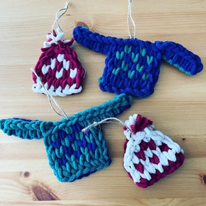May include: Four handmade knitted ornaments in the shape of hats and sweaters. Two of the ornaments are red and white, one is blue and green, and one is teal and green.