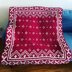 Pode incluir: Uma manta de malha vermelha e branca com um padrão geométrico. A manta tem uma borda branca com um padrão repetitivo de triângulos e diamantes.