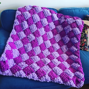 May include: A purple and lavender checkered knit blanket with a fluffy texture. The blanket is draped over a blue couch.