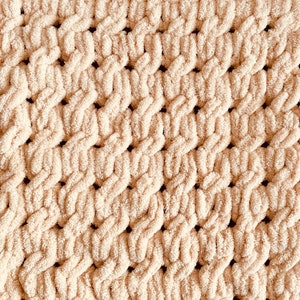 May include: A close-up of a cream-colored knitted blanket with a textured, woven pattern. The blanket is made of soft, fluffy yarn and has a subtle sheen.