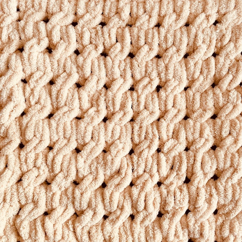 May include: A close-up of a cream-colored knitted blanket with a textured, woven pattern. The blanket is made of soft, fluffy yarn and has a subtle sheen.