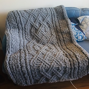 Puede incluir: Una manta de punto grueso de color gris con un patrón de diamantes. La manta está colocada sobre un sofá.