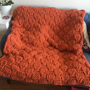 May include: A large, orange knitted throw blanket with a textured, woven pattern. The blanket is folded and draped over a couch.