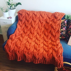 Könnte beinhalten: Eine dicke Strickdecke in verbrannter Orange mit einem Zopfmuster. Die Decke liegt über einem blauen Sofa.