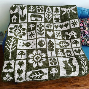May include: A large, olive green and white knitted blanket with a repeating pattern of nature-themed images, including trees, flowers, and animals. The blanket is draped over a blue couch, showcasing its intricate design and cozy texture.