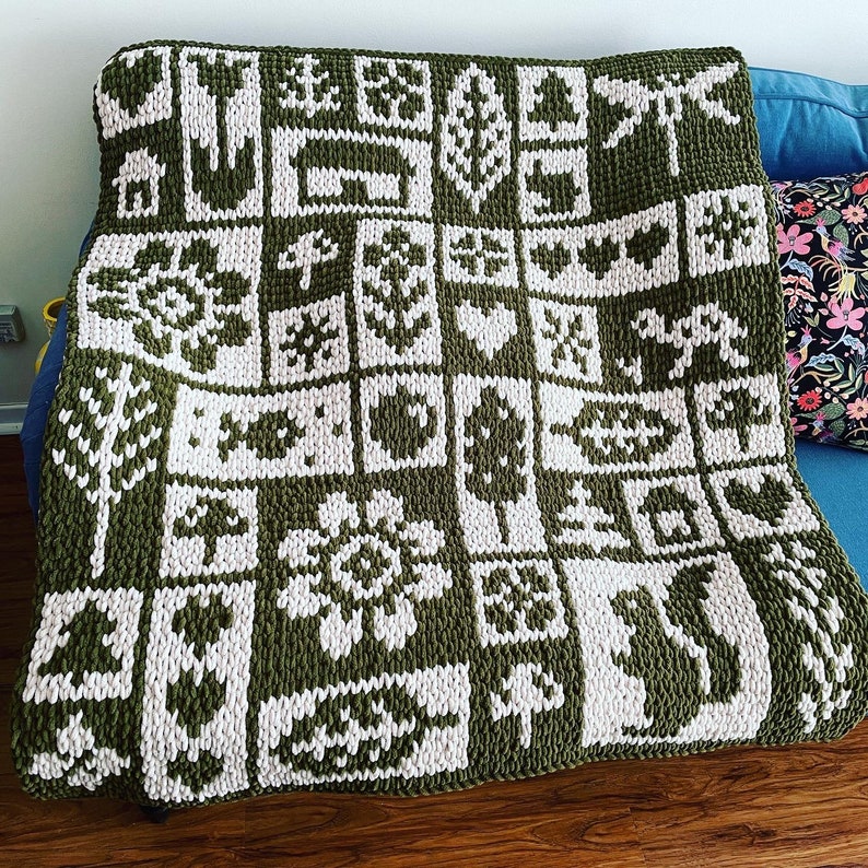 May include: A large, olive green and white knitted blanket with a repeating pattern of nature-themed images, including trees, flowers, and animals. The blanket is draped over a blue couch, showcasing its intricate design and cozy texture.