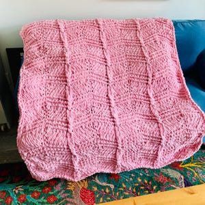 May include: A light pink chunky knit blanket featuring a repeating textured pattern of vertical columns and diamond-shaped stitches. The throw is crafted from thick, soft yarn with a consistent pastel pink colour throughout the entire surface.
