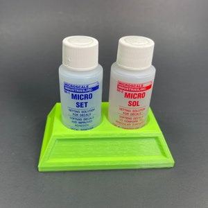 May include: Two small bottles of Micro Set and Micro Sol decal setting solutions for model building. The bottles are on a green plastic stand. The bottles are labeled with the Micro Scale Industries logo and product names.
