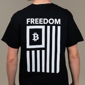 May include: Black t-shirt with a white graphic on the back. The graphic features the word "FREEDOM" above a Bitcoin symbol within a white square, and white vertical stripes resembling a flag. The shirt is worn by a person.