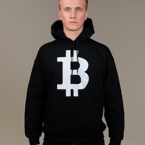 May include: Black hooded sweatshirt with a large white Bitcoin symbol on the front. The sweatshirt has a kangaroo pocket and a drawstring hood. The person is wearing black jeans.
