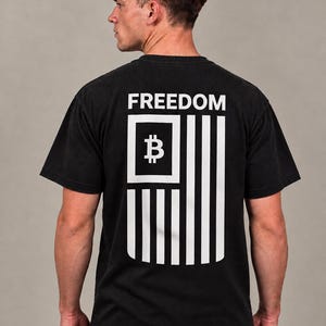 May include: A black short-sleeve t-shirt featuring a white graphic on the back. The design displays the word FREEDOM above a stylized flag motif where the canton contains a Bitcoin symbol, followed by vertical stripes representing the flag design.