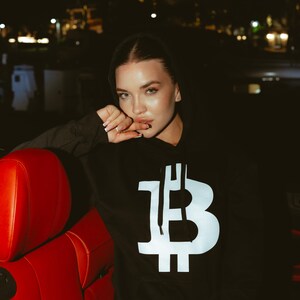 May include: A black hooded sweatshirt featuring a large, white, centered Bitcoin symbol graphic on the front. The hoodie includes a drawstring hood and a front kangaroo pocket, constructed from a solid black fabric with a bold, high-contrast design.