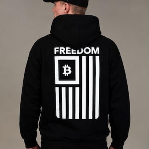 May include: A black hooded sweatshirt featuring a white graphic on the back. The graphic displays the word FREEDOM above a stylized flag design containing a Bitcoin symbol inside a square. A black baseball cap with white text is also worn.
