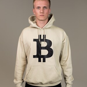 May include: Beige hoodie with a large black Bitcoin symbol on the front. The hoodie has a kangaroo pocket and a drawstring hood. The person in the image is wearing black pants.