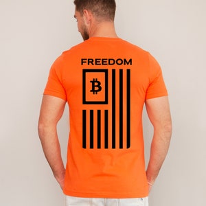 May include: Orange t-shirt with a black graphic of an American flag design. The flag features a Bitcoin symbol in the upper left corner and the word "FREEDOM" above the flag.