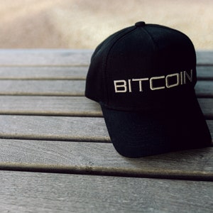 May include: A black baseball cap featuring the word BITCOIN embroidered in a light-colored, sans-serif font across the front panel. The cap has a structured crown, a curved brim, and a button at the top center, showcasing a clean and simple design.