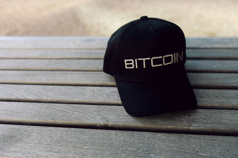 May include: A black baseball cap featuring the word BITCOIN embroidered in a light-colored, sans-serif font across the front panel. The cap has a structured crown, a curved brim, and a button at the top center, showcasing a clean and simple design.