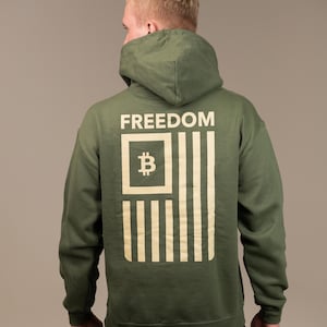 May include: Olive green hoodie with the word "FREEDOM" printed across the upper back. Below the word is a graphic resembling the American flag, with a Bitcoin symbol in the upper left corner. The hoodie is made of a soft material.