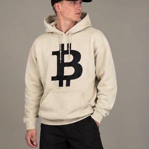 May include: A beige hooded sweatshirt featuring a large black Bitcoin symbol on the chest, paired with black cargo pants and a black baseball cap with a white Bitcoin logo and the Roman numeral IX embroidered on the side panel.