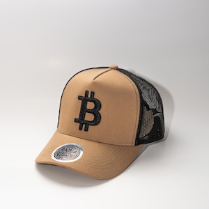 May include: Tan trucker hat with a black mesh back and a black Bitcoin symbol embroidered on the front. The hat has a curved brim and a snapback closure. A sticker is on the brim.