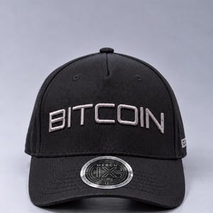 May include: A black baseball cap featuring the word BITCOIN embroidered in light gray thread across the front panels. A circular silver and black sticker is affixed to the center of the curved brim, displaying the text MERCH and Est. 2019.
