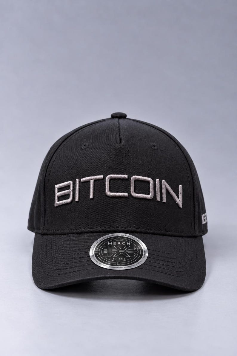 May include: A black baseball cap featuring the word BITCOIN embroidered in light gray thread across the front panels. A circular silver and black sticker is affixed to the center of the curved brim, displaying the text MERCH and Est. 2019.