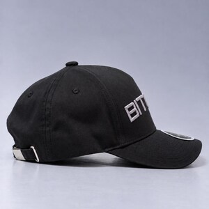 May include: A black baseball cap featuring the text BIT embroidered in white on the front panel. The cap has a curved brim, a top button, and a silver-toned metal adjustable buckle strap at the back. A small circular sticker is visible on the brim.