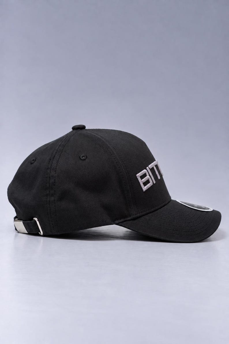 May include: A black baseball cap featuring the text BIT embroidered in white on the front panel. The cap has a curved brim, a top button, and a silver-toned metal adjustable buckle strap at the back. A small circular sticker is visible on the brim.