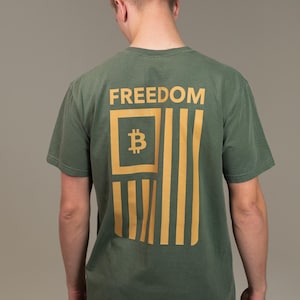 May include: Olive green t-shirt with the word "FREEDOM" above a Bitcoin-themed flag design in gold. The flag features a Bitcoin symbol within a square and vertical stripes. The shirt is a short-sleeved crew neck.