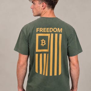 May include: A short-sleeve olive green t-shirt featuring a large mustard yellow graphic on the back. The graphic depicts a stylized flag design with the Bitcoin symbol in the canton and the word FREEDOM printed in bold capital letters above it.
