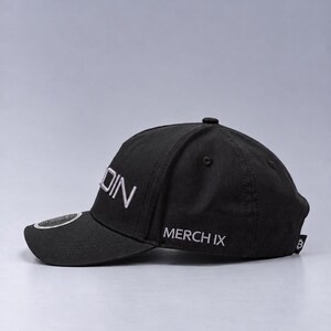 May include: A black baseball cap featuring the white embroidered text DIN on the side panel and MERCH IX on the lower side. The cap includes a small circular emblem on the brim and a small black tag with a white Bitcoin symbol on the back edge.