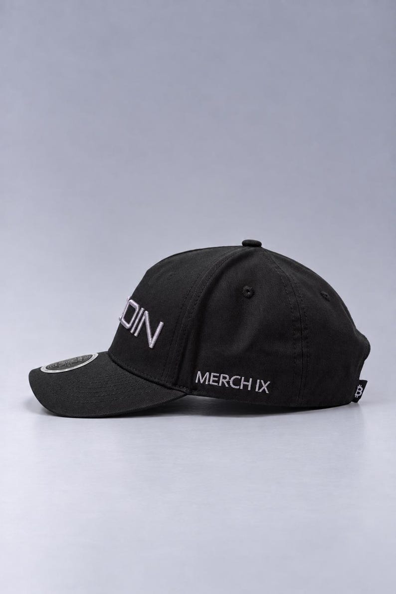 May include: A black baseball cap featuring the white embroidered text DIN on the side panel and MERCH IX on the lower side. The cap includes a small circular emblem on the brim and a small black tag with a white Bitcoin symbol on the back edge.