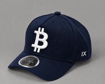 May include: A navy blue baseball cap featuring a large, white, embroidered Bitcoin symbol on the front panel. The side of the cap displays the Roman numeral IX in white embroidery, and a circular silver sticker is on the brim.