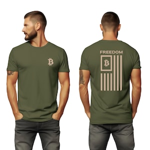 May include: Olive green t-shirt featuring a Bitcoin symbol on the front and a stylized American flag design with the word "FREEDOM" on the back. The flag design incorporates the Bitcoin symbol within its stripes.