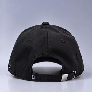 May include: A black baseball cap viewed from the back, featuring a fabric strap with a silver-toned metal buckle. The strap displays a small white Bitcoin symbol, and the side of the cap shows partial white text that reads IX.