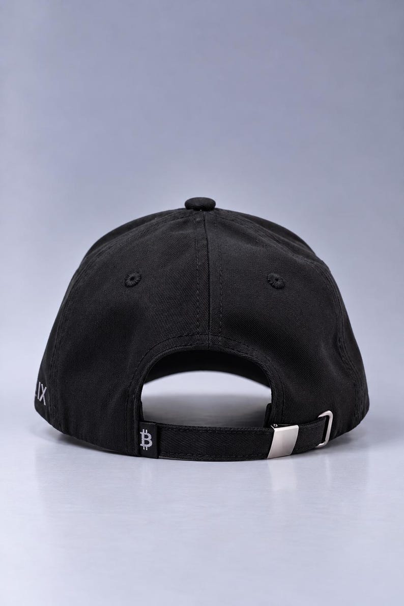 May include: A black baseball cap viewed from the back, featuring a fabric strap with a silver-toned metal buckle. The strap displays a small white Bitcoin symbol, and the side of the cap shows partial white text that reads IX.